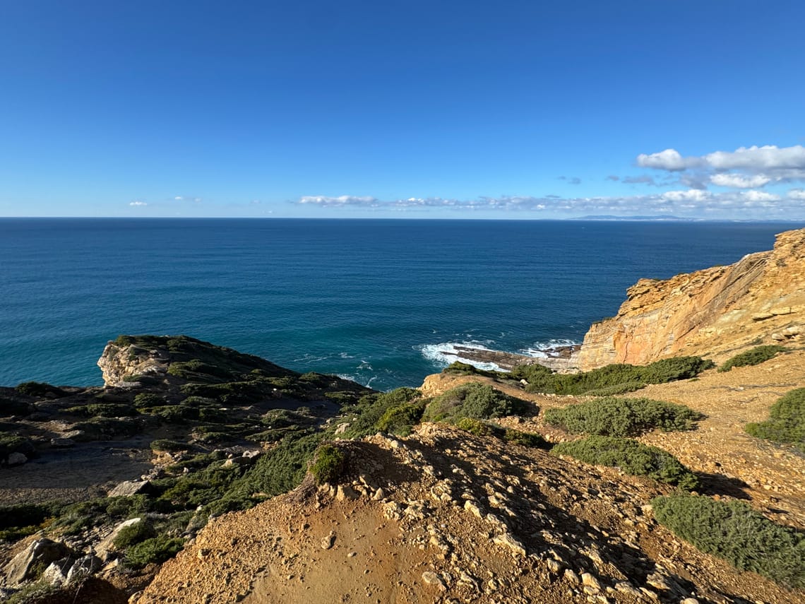 Yellow cliffs facing a dark blue ocean under a bright blue sky. On the horizon, we see the hills of Sintra.